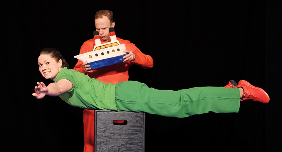 En kvinna i grön dräkt ligger på en piedestal med armarna sträckta rakt ut. Bakom henne står en man med en leksaksbåt i handen.