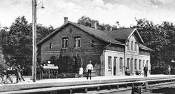 Bild på Vårgårda järnvägsstation från 1920. Folk promenerar framför en äldre byggnad, Bilden är svartvit.